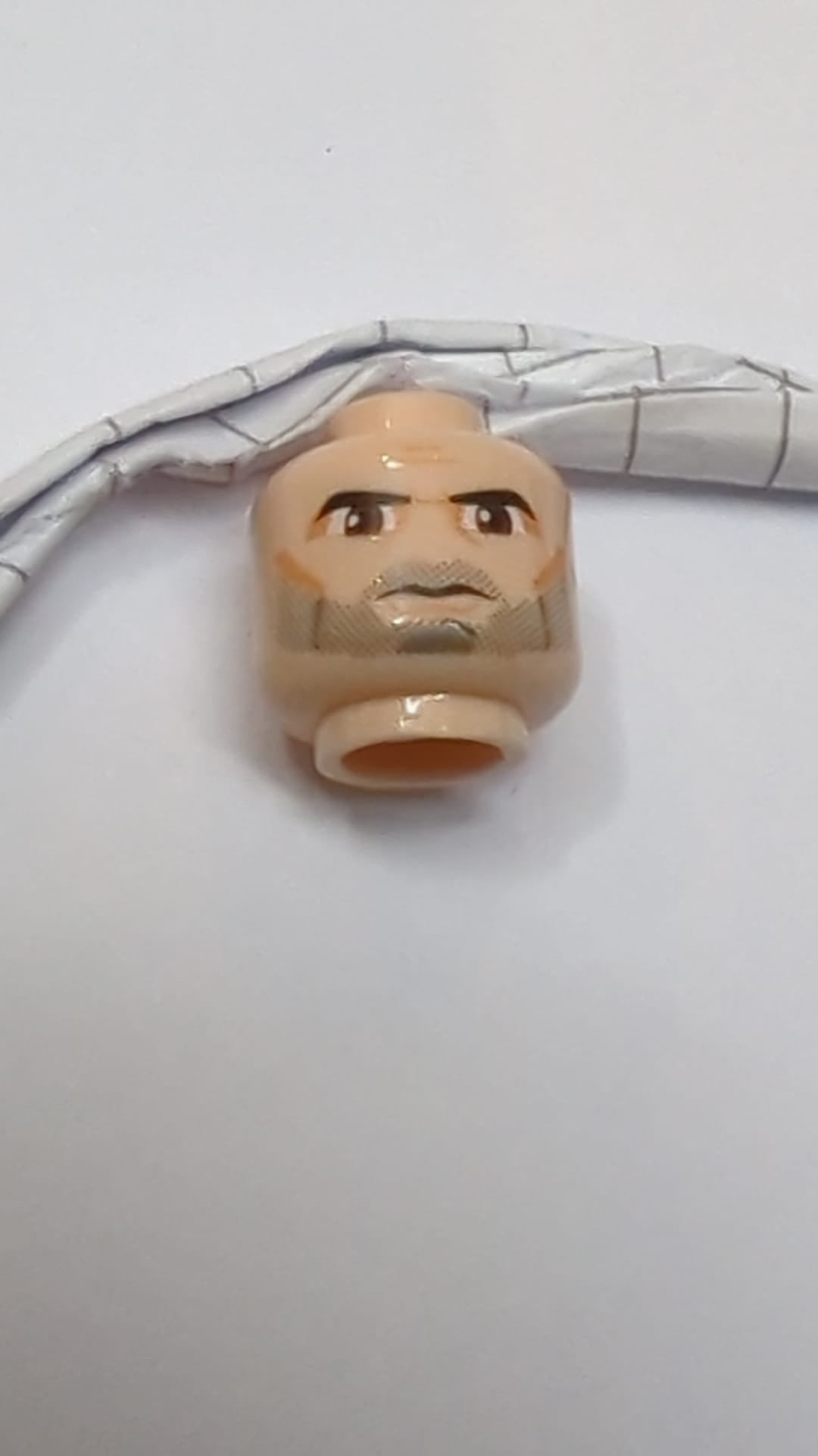 Minifigur, Kopf: Schwarze, dichte Augenbrauen und Bartstoppeln, große, rotbraune Augen, nougatfarbener Lidschatten, Wangenfalten und Grübchen am Kinn, zusammengezogene Stirn, strenges Gesichtsmuster hellnougat (fleischfarben)