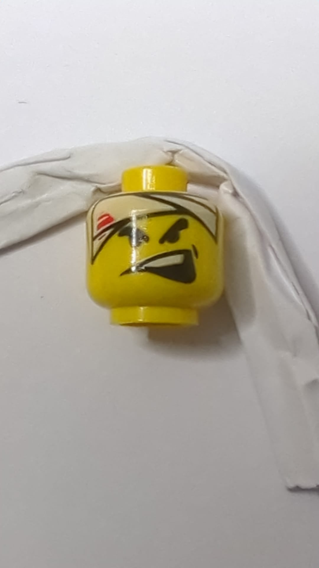 Minifigur, Kopf: Schwarze, dicke Augenbrauen, weißer Kopfverband mit roten Streifen, Grübchen, weit geöffneter, schiefer Mund mit sichtbaren oberen Zähnen gelb