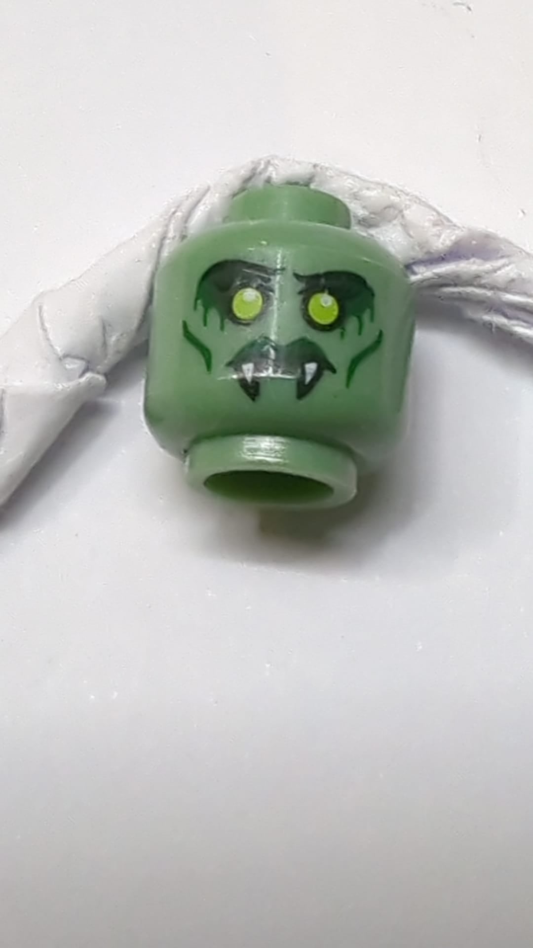 Minifigur, doppelseitiger Alienkopf mit gelben Augen, Reißzähnen, grünen Lippen, Wangenfalten / offenem Mund mit rotem Augenmuster sandgrün