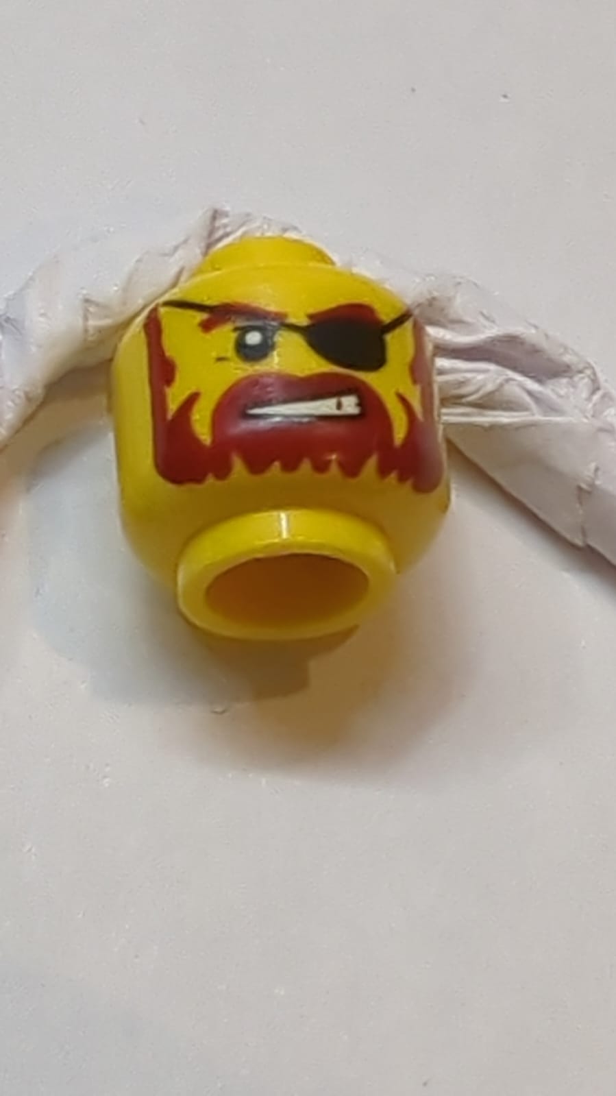 Kopf mit dunkelroten Augenbrauen, Schnurrbart und Vollbart, schwarzes Augenpflaster, schiefes offenes Mundlächeln mit Zahnabdruck