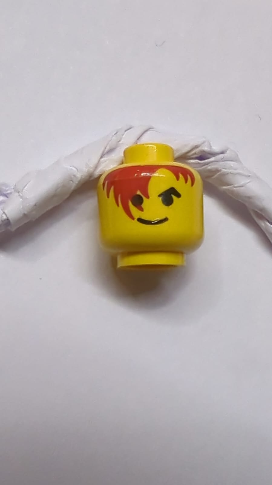 Kopf mit schwarzen, schrägen Augenbrauen, dunkelrotem Haar, das über das rechte Auge fällt, standardmäßiges Grinsen, gelb