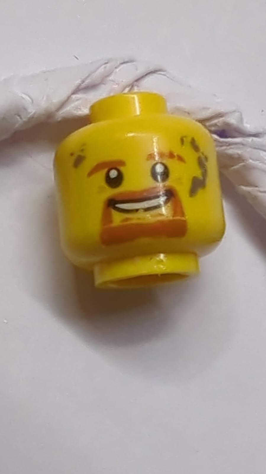 Kopf dunkelorangefarbene Augenbrauen, Kinnbart und Schnurrbart, dunkelbläulich-graue Flecken, offen lächelnder Mund mit Zahnmustern gelb