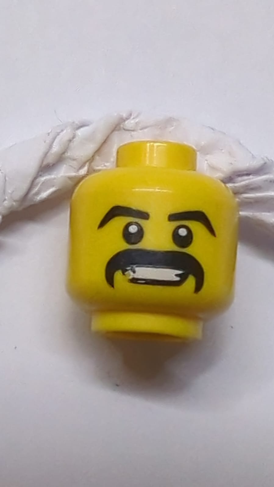 Kopf mit schwarzen, schrägen Augenbrauen und dickem Schnurrbart, offener Mund mit sichtbaren Zähnen beim Lächeln gelb