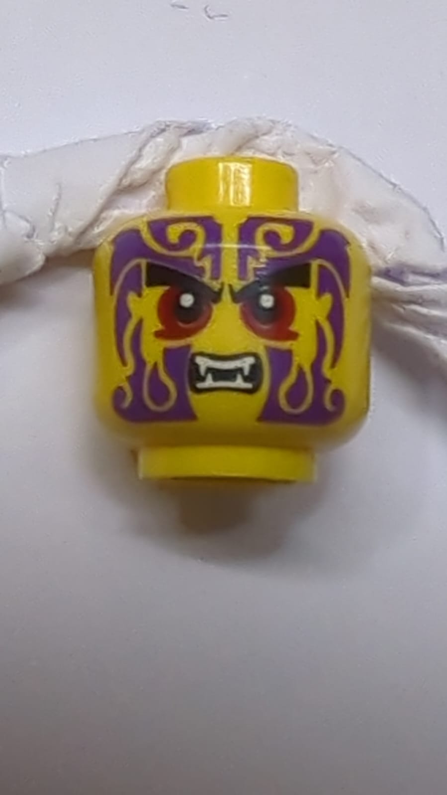 Kopf mit schwarzen Augenbrauen, dunkelroten Augen, dunkelvioletten Tattoos, offenem Mund mit Reißzahn-Muster gelb