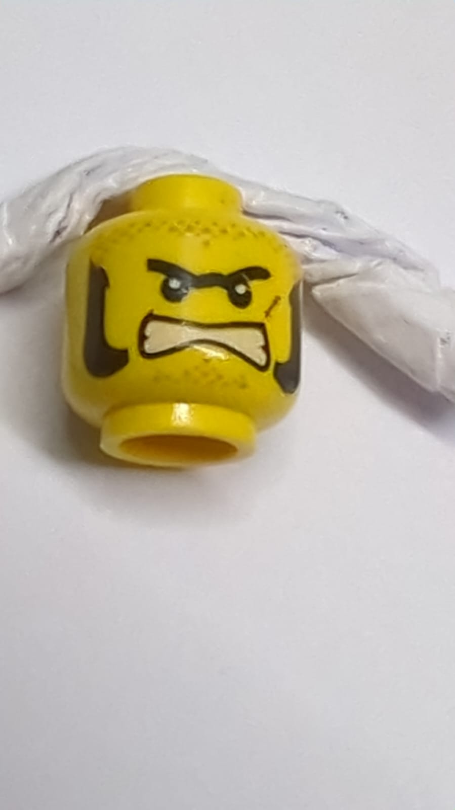 Minifigur, Kopf mit beidseitigem schwarzen, dicken Monobrauen- und Kotelettenmuster, dunkelbraunen Schmutzflecken, rötlich-brauner Narbe, wütend gefletschten Zähnen / dunkelblau-graues Bandana-Muster über dem Mund gelb