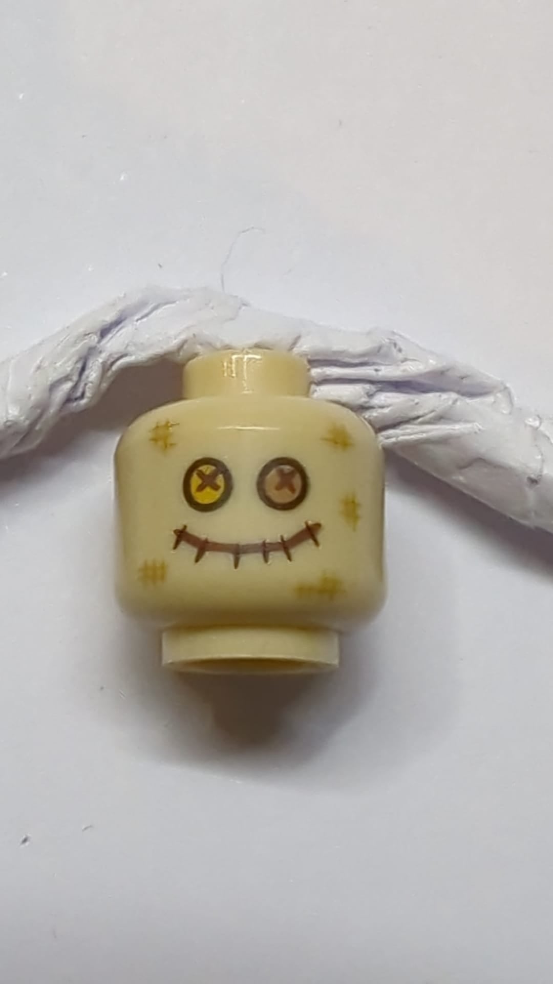 Minifigur, Kopf mit dunkelbraunen Abriebspuren, hellorange und mittelbraune Knopfaugen, genähter Mund mit Vogelscheuchenmuster beige