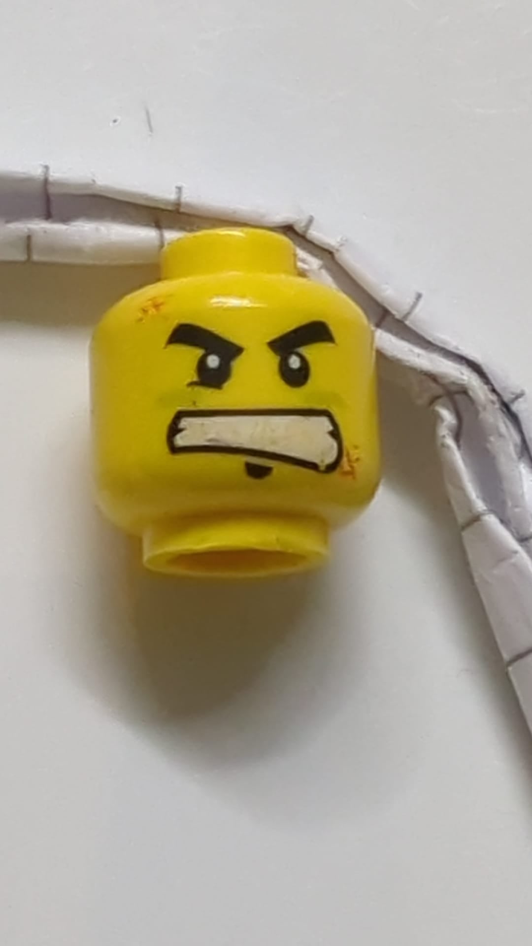 Minifigur, Kopf mit doppelseitiger schwarzer, dicker Augenbrauenpartie und Kinnbart, rechtes Unterlid, limettengrüne Farbe unter den Augen, orangefarbene Abriebspuren, strenger Gesichtsausdruck mit Grübchen/wütend entblößten Zähnen gelb