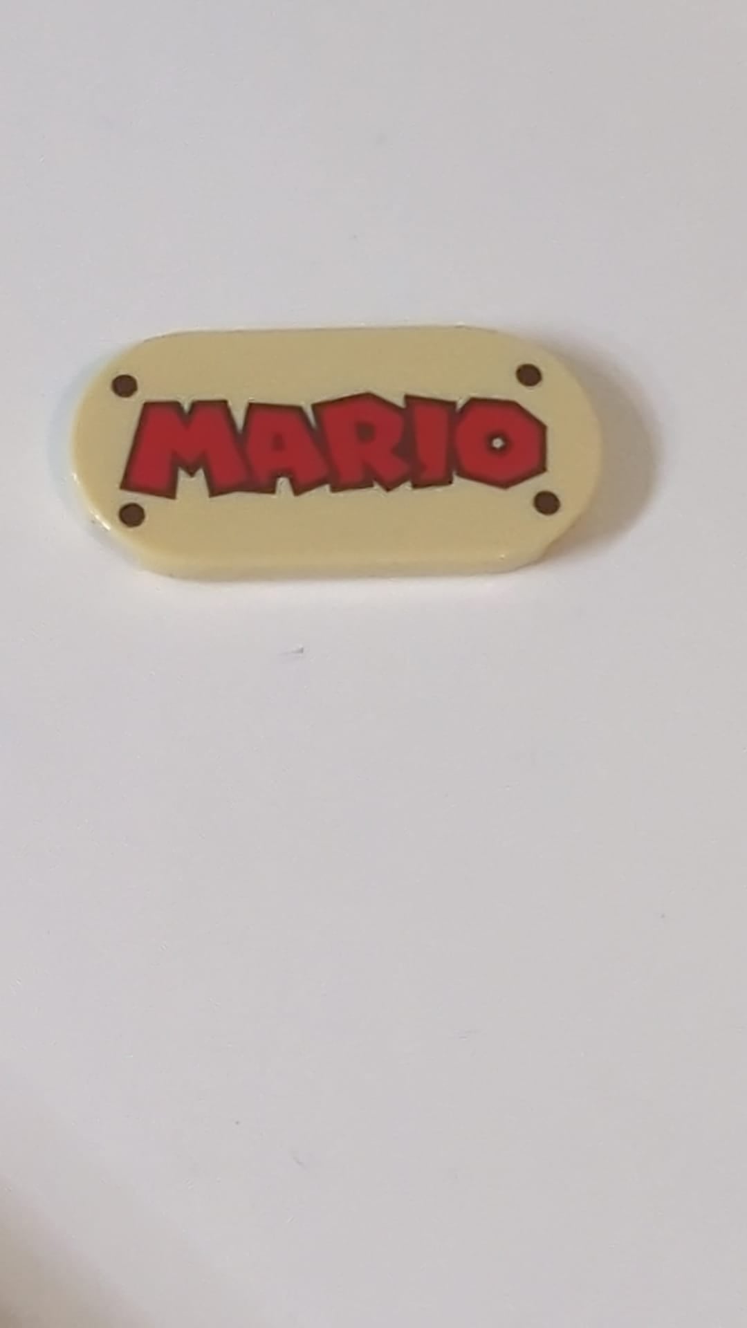 2x4 Fliese, rund (oval) mit rotem „MARIO“-Schriftzug und dunkelbraunem Nietmuster beige