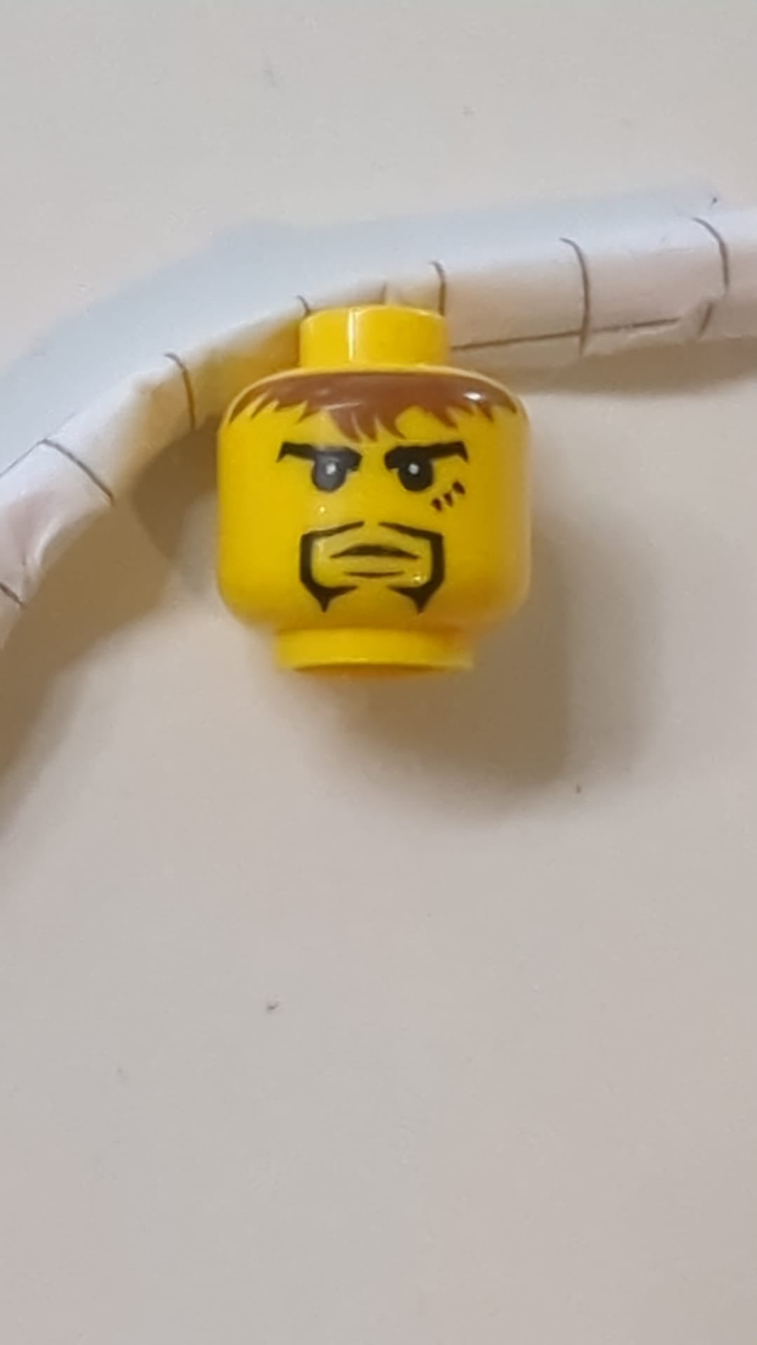 Minifigur, Kopf: Schwarzer Spitzbart, wuscheliges braunes Haar, gespaltenes Kinn, 3 Markierungen unter dem linken Auge, Muster: „Adric“ gelb