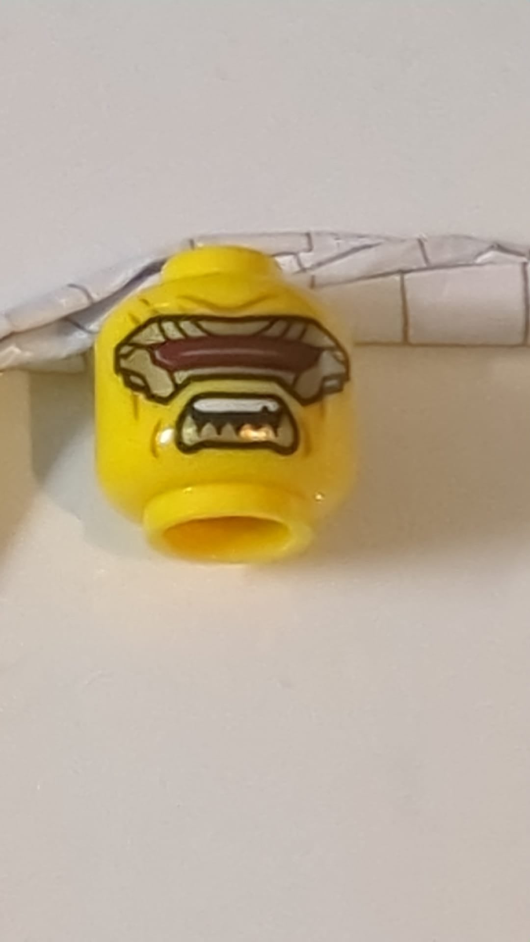 Minifigur, Kopf: Doppelseitige goldene Augenmaske mit dunkelrotem Visier, mittelbraune Wangenfalten, neutraler/offener Mund mit spitzen unteren Zähnen gelb