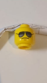 Minifigur, Kopf: Schwarze Sonnenbrille mit dunkelsilbernen Reflexen, mittelgroßes nougatfarbenes Grübchen am Kinn, grimmiges Grinsen gelb