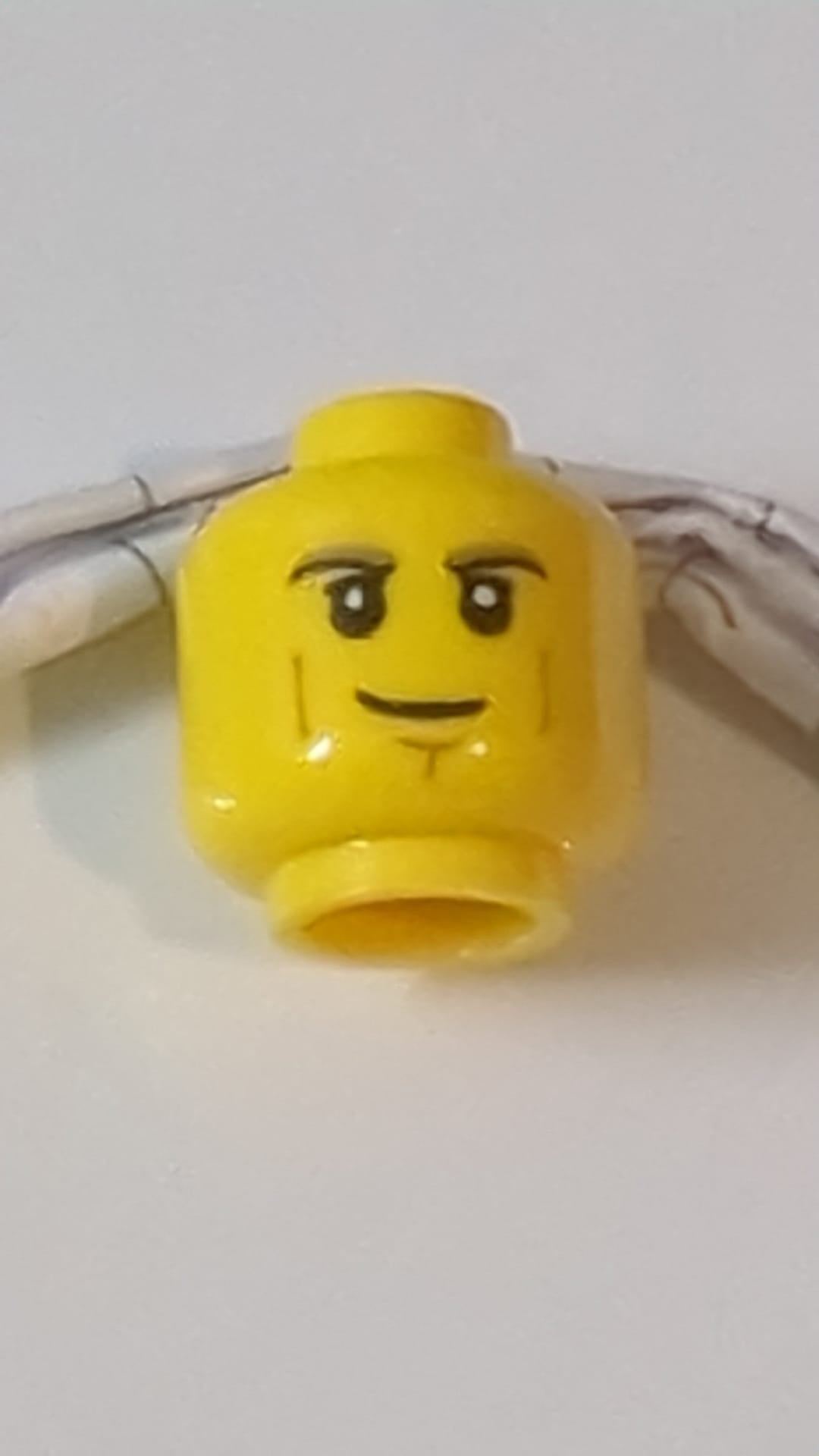 Minifigur, Kopf: Dunkelbraun mit schwarzen Augenbrauen und Oberlidern, mittelbraune Wangenfalten und Grübchen am Kinn, asymmetrisches Grinsen gelb
