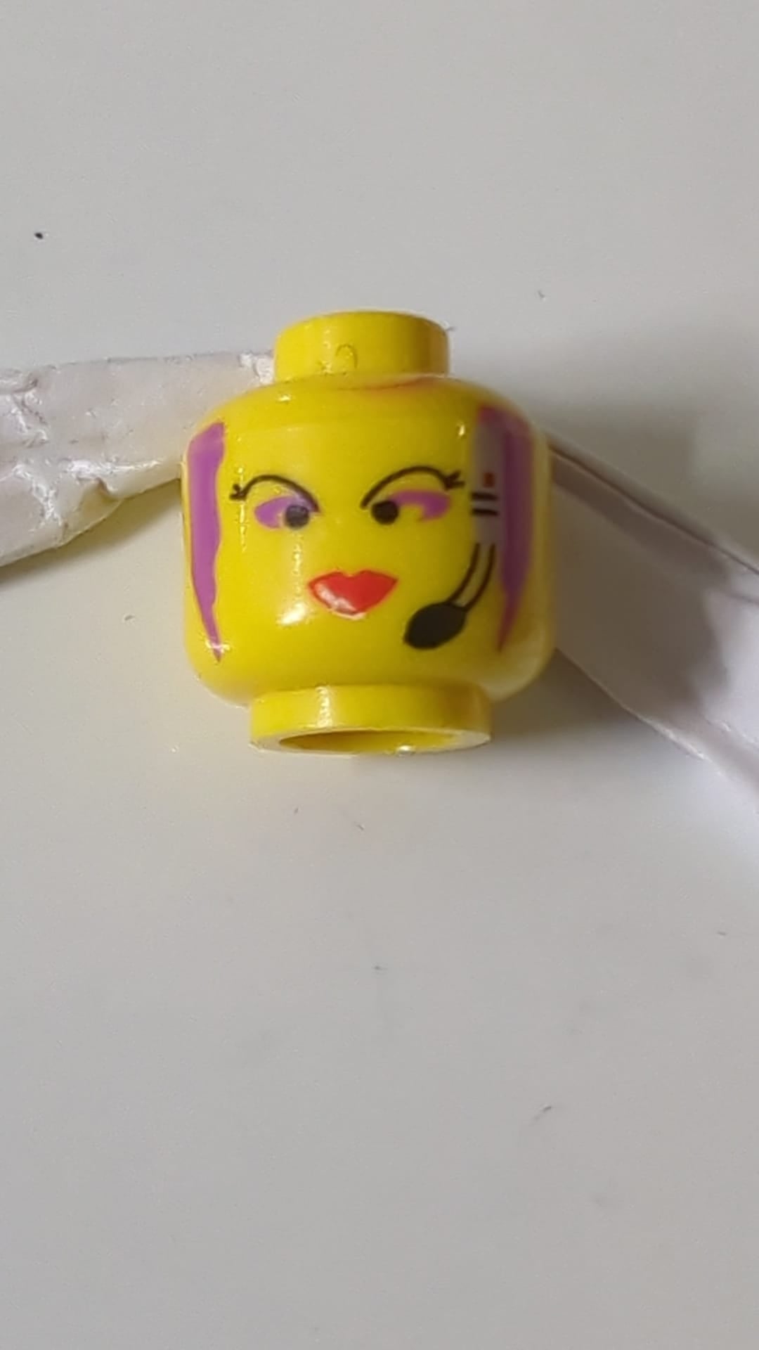 Minifigur, weiblicher Kopf mit dünnen schwarzen Augenbrauen, lila Haaren und Lidschatten, Headset mit Mikrofon und schmalen roten Lippen. gelb