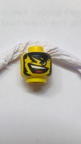 Kopf mit schwarzen buschigen Augenbrauen, Haaren und Koteletten, Augenzwinkern, schiefes offenes Mundlächeln mit oberen Zähnen und roter Zunge gelb