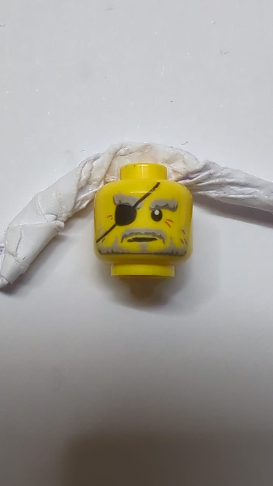 Grauer Kopf, graue Augenbrauen und Bart, schwarzes Augenpflaster, dunkelorange Krähenfußmuster gelb