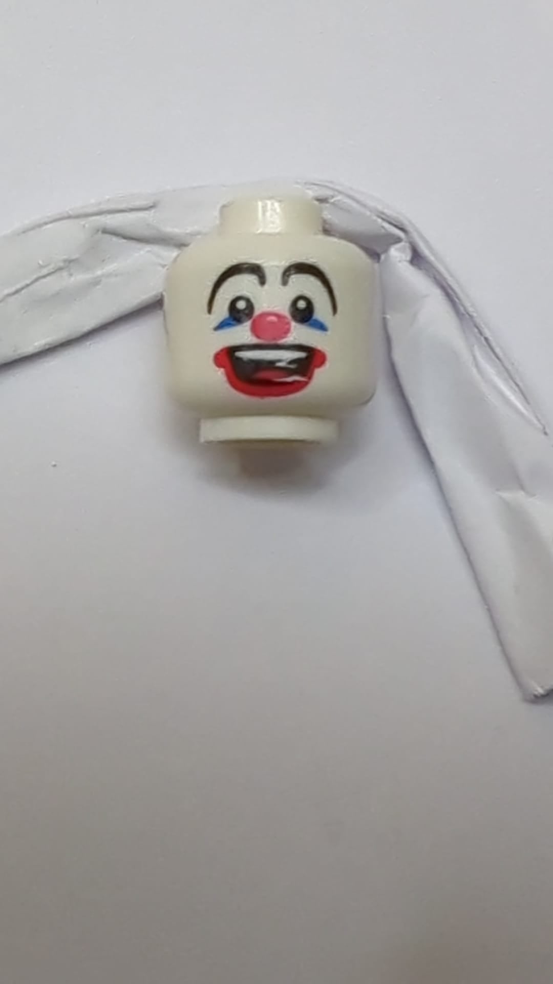 Minifigur, Kopf: Schwarze, lange, geschwungene Augenbrauen, blauer Lidschatten, rote Clownsnase und aufgemalte Lippen, breites, offenes Lächeln mit sichtbaren oberen Zähnen und Zungenmuster weiß