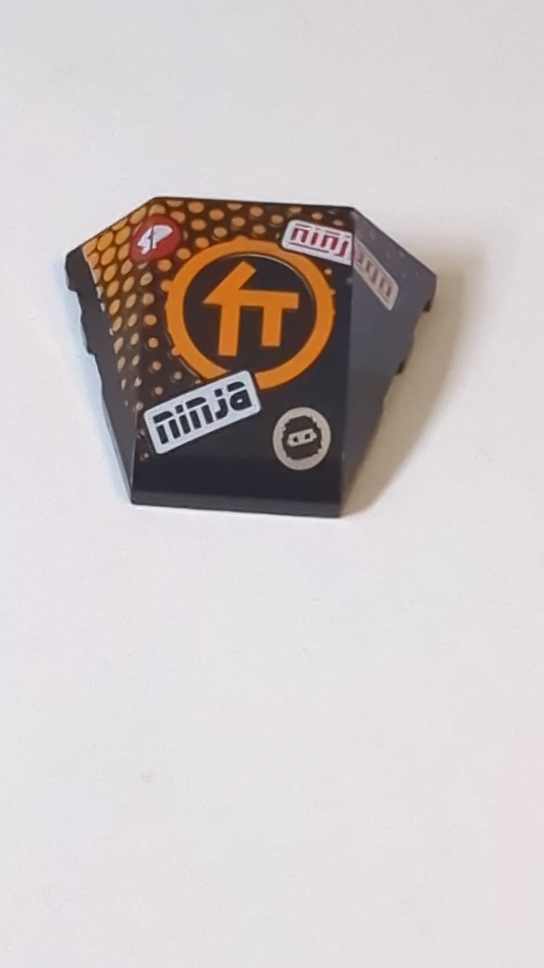 4x3 Keil Dreifach gebogen, ohne Noppen, mit orangefarbenem Ninjago-C-Symbol im Kreis, orangefarbenen Punkten, „ninja“ und „ninjago“-Muster schwarz