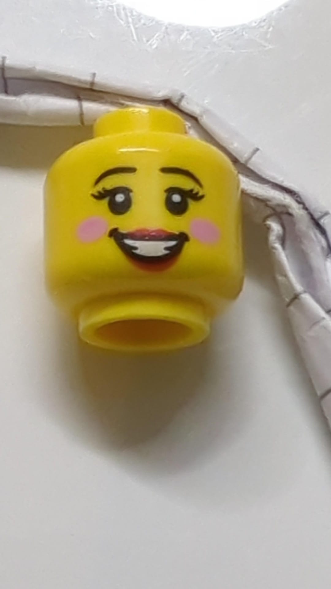 Minifigur, weiblicher Kopf: schwarze Augenbrauen, Wimpern, leuchtend pinkfarbene Kreise auf den Wangen, rote Lippen, offenes Lächeln mit Zahnmuster gelb