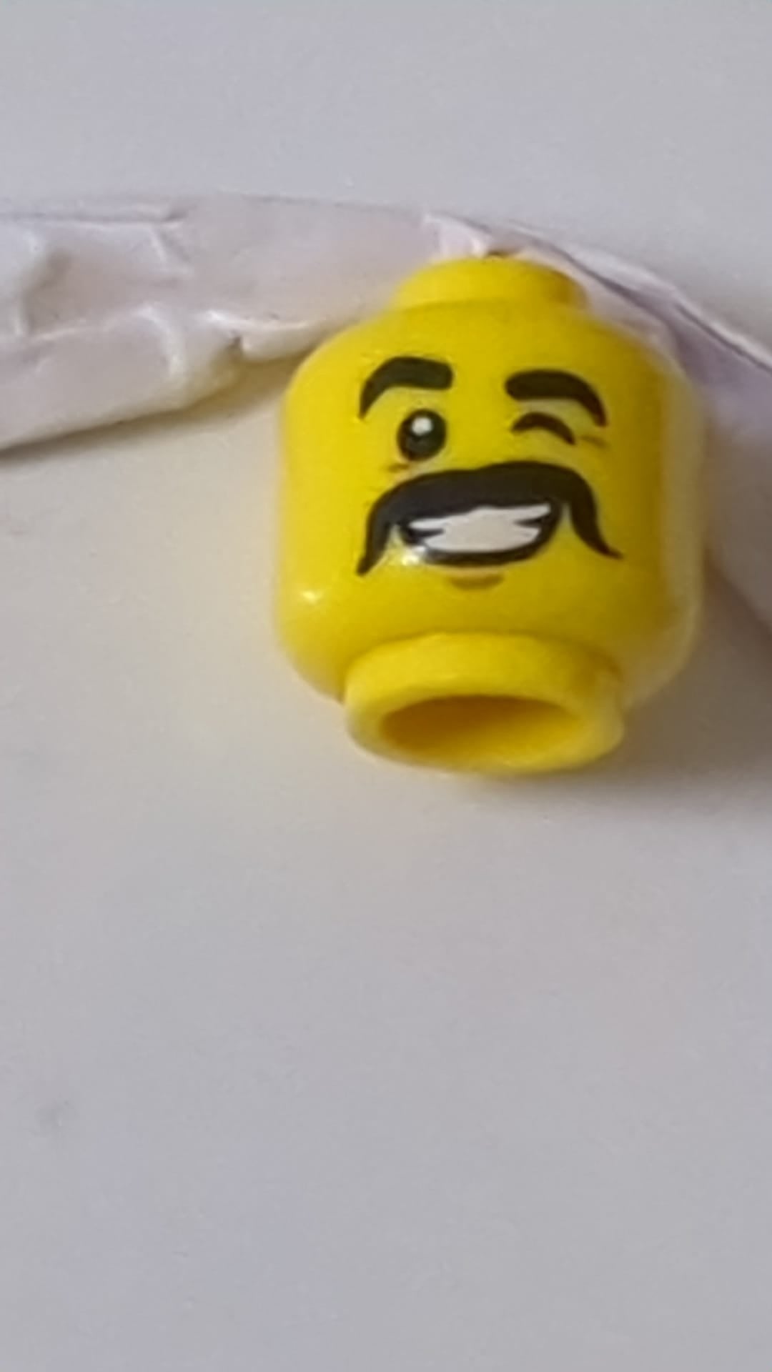 Minifigur, männlicher Kopf mit dicken schwarzen Augenbrauen und Schnurrbart, zwinkert, lächelt mit weißem Zahnmuster gelb