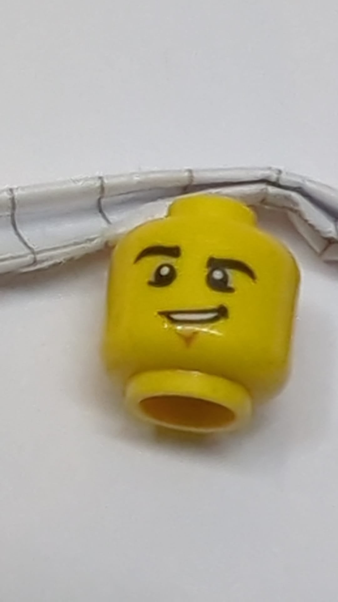 Minifigur, männlicher Kopf, schwarze Augenbrauen, hochgezogene rechte Augenbraue, Grübchen am Kinn und schiefes Grinsen mit Zahnmuster gelb