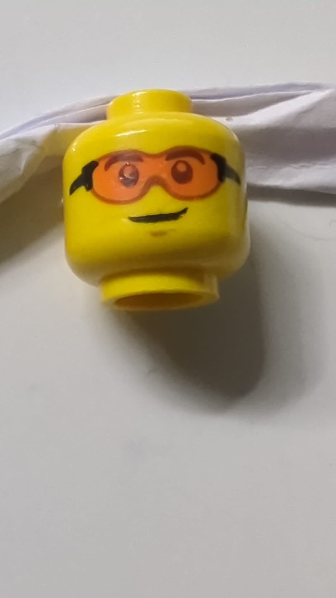 Minifigur, Schutzbrille mit orangefarbenen Gläsern und schwarzem Gestell, Grübchen am Kinn, schiefes Grinsen gelb