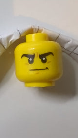 Minifigur, Kopf mit doppelseitigen, dicken schwarzen Augenbrauen, mittelgroßem nougatfarbenem Grübchen am Kinn, entschlossenem Grinsen / finsterem Blick mit geöffnetem Mund und leicht geöffneten Zähnen gelb
